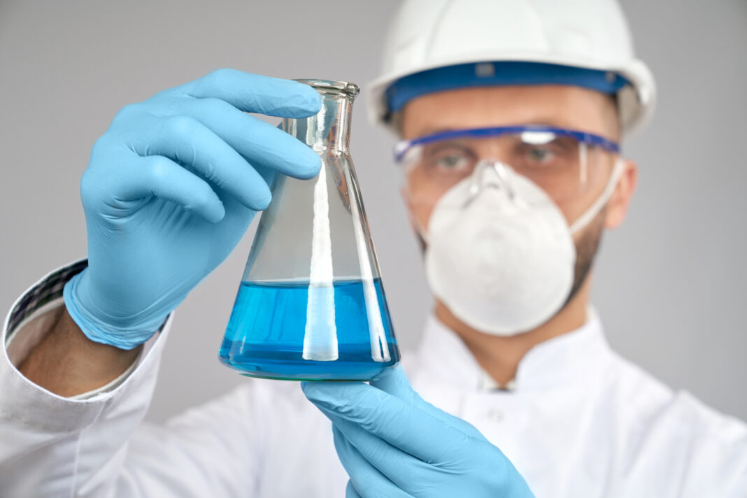 Close up of young chemist in glasses, helmet and mask doing experiment in laboratory, isolated on grey. Selective focus of flask with blue liquid in hand of scientist in gloves and white lab coat.